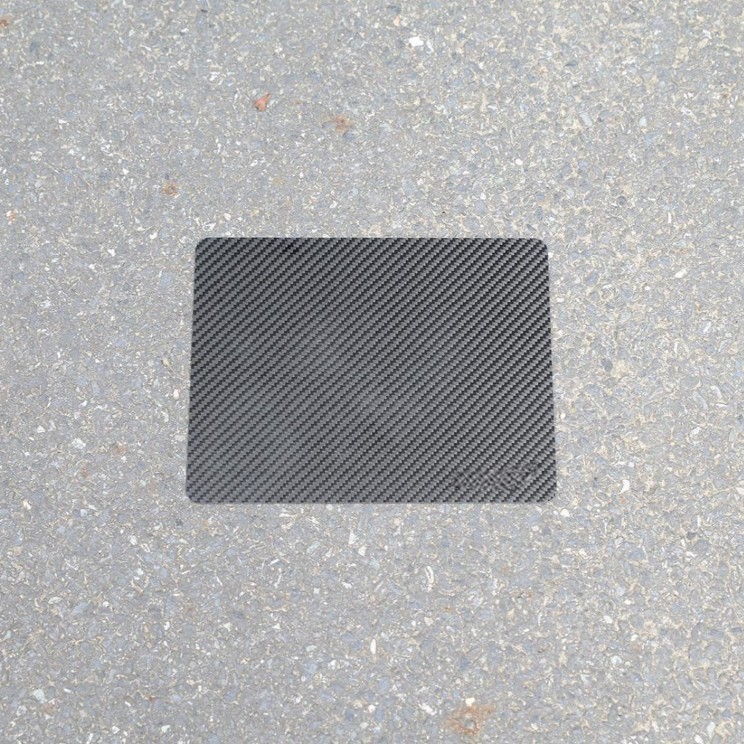 "A rectangular 3K carbon fiber mouse pad placed on a rough asphalt surface."