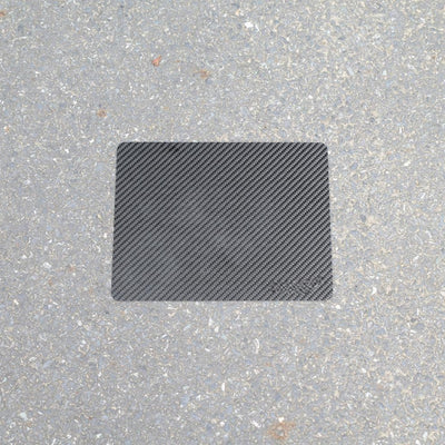 A rectangular 3K carbon fiber mouse pad placed on a rough asphalt surface