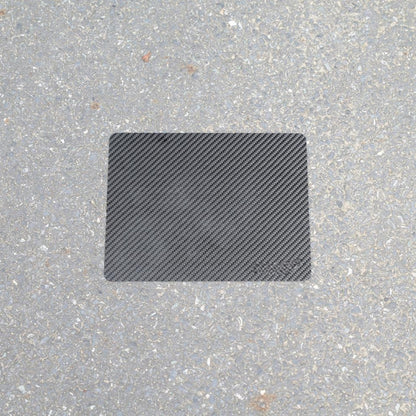 "A rectangular 3K carbon fiber mouse pad placed on a rough asphalt surface."