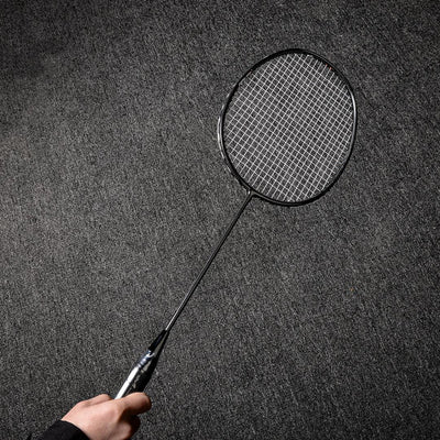 Close-up of a carbon fiber badminton racket with a distinct texture, held by a hand, against a gray textured background.