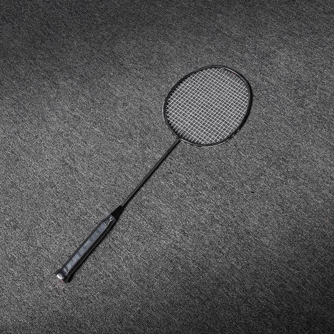Close-up of a carbon fiber badminton racket with a black frame, shaft, and textured grip, laid on a gray textured surface.