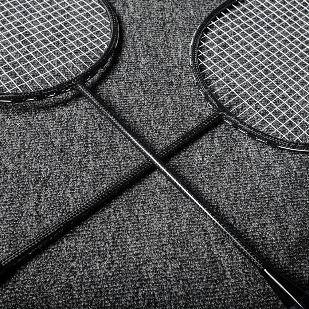 Close-up of two carbon fiber badminton rackets with black frames and textured shafts, crossed on a gray textured surface.