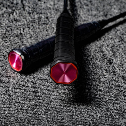 Close-up of the handles of two carbon fiber badminton rackets, featuring black grips and pink octagonal end caps with a textured finish, placed on a gray textured surface.