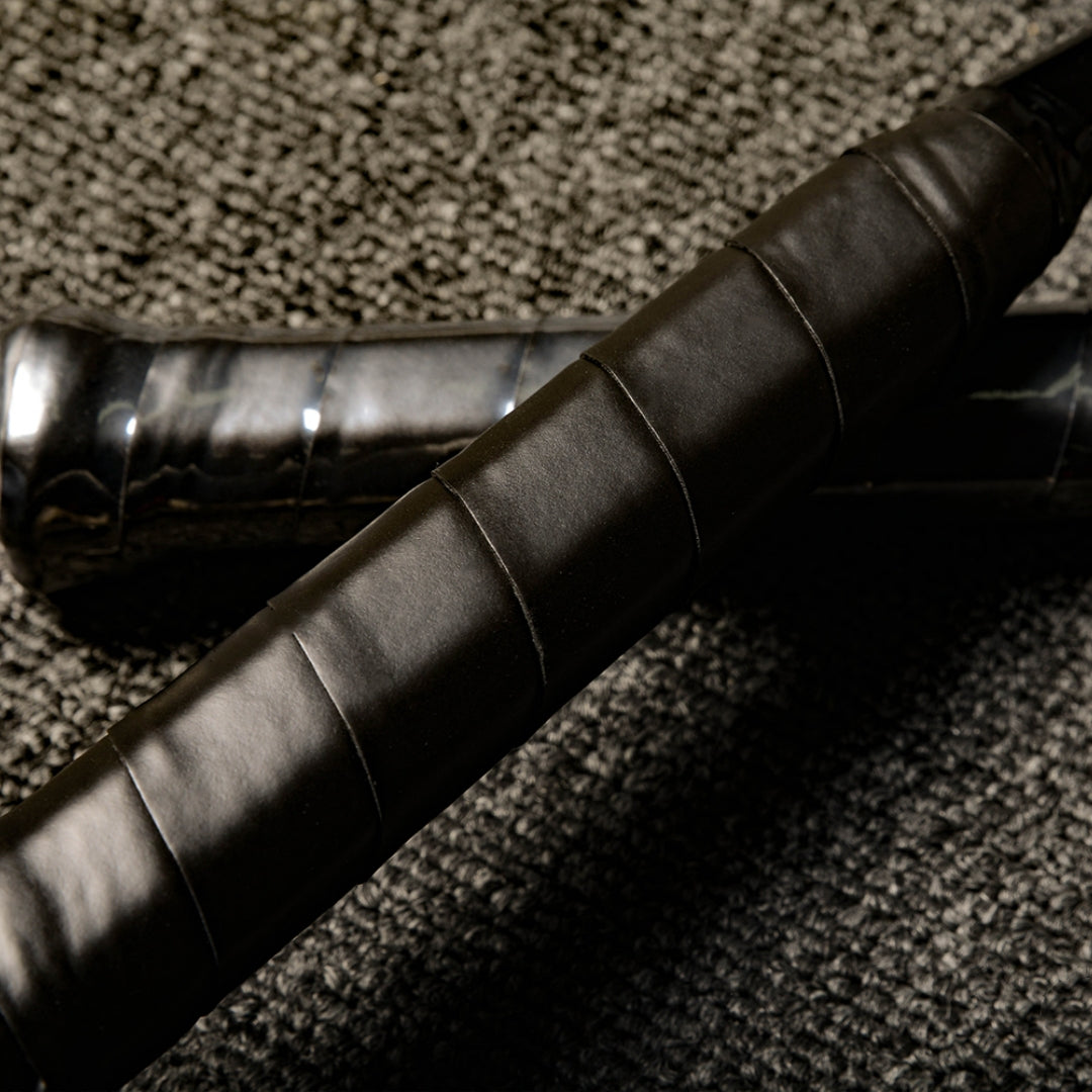 Close-up of the handles of two carbon fiber badminton rackets with black textured grips, laid on a gray textured surface.