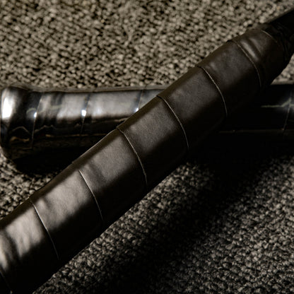 Close-up of the handles of two carbon fiber badminton rackets with black textured grips, laid on a gray textured surface.