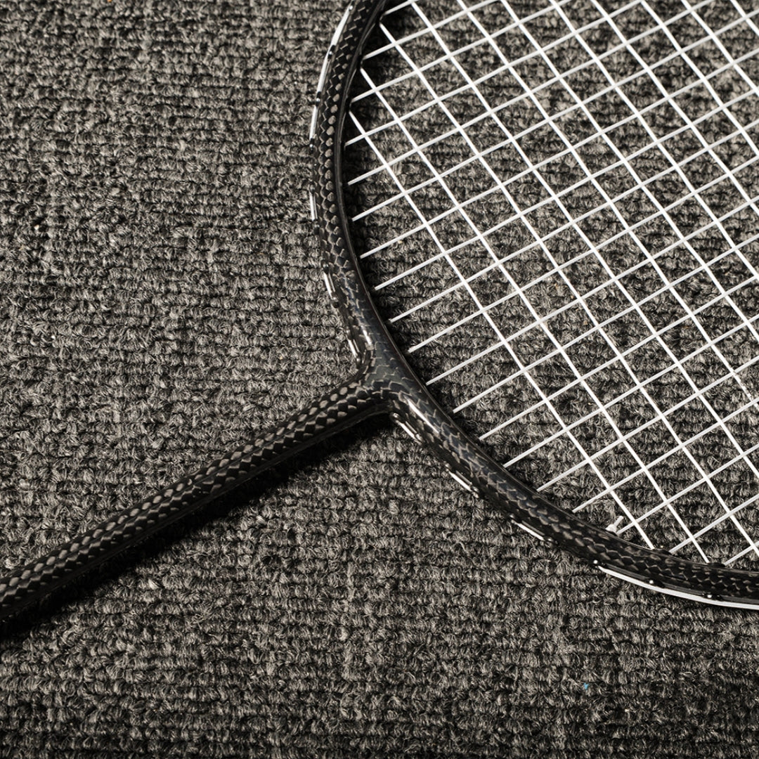 Close-up of a carbon fiber badminton racket showcasing the distinct carbon fiber texture on the frame and shaft, with white strings, laid on a gray textured surface.