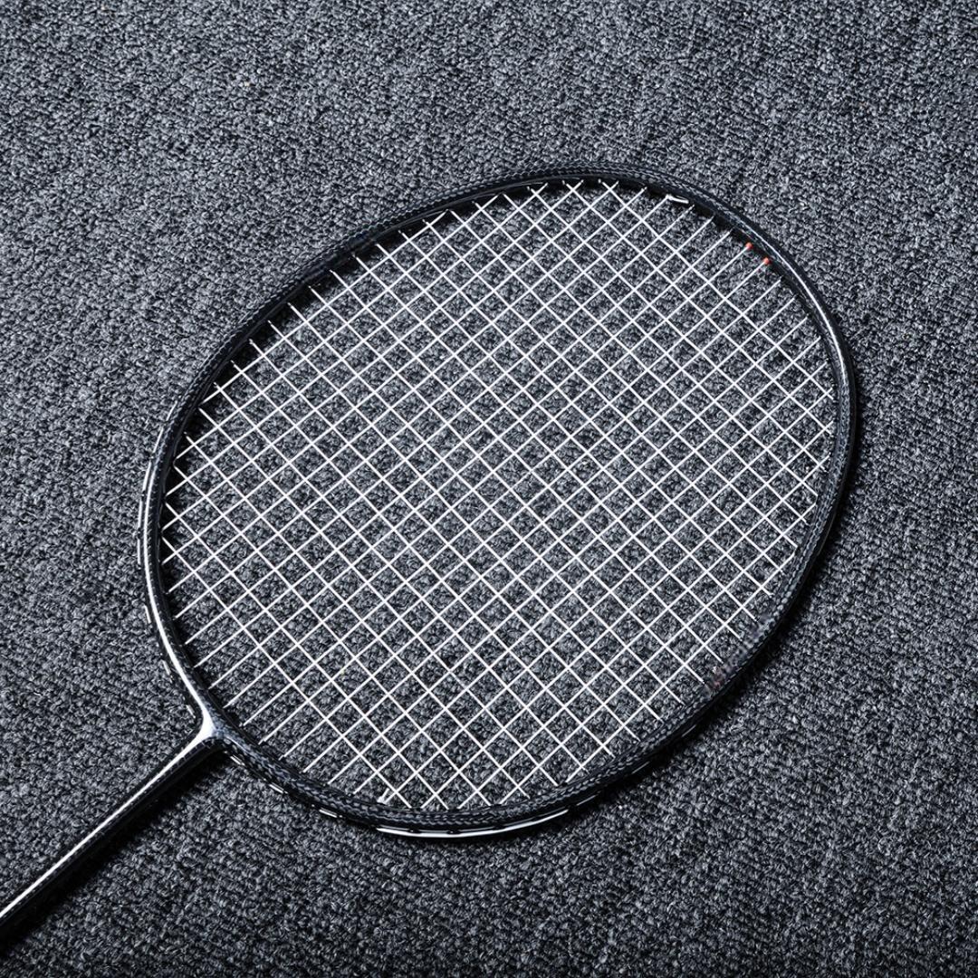 Close-up of a carbon fiber badminton racket with a black frame and white strings, laid on a gray textured surface.