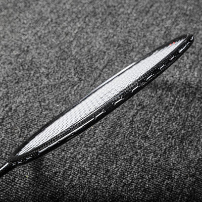 Close-up of a carbon fiber badminton racket frame showcasing its textured surface and white strings, positioned on a gray textured background.