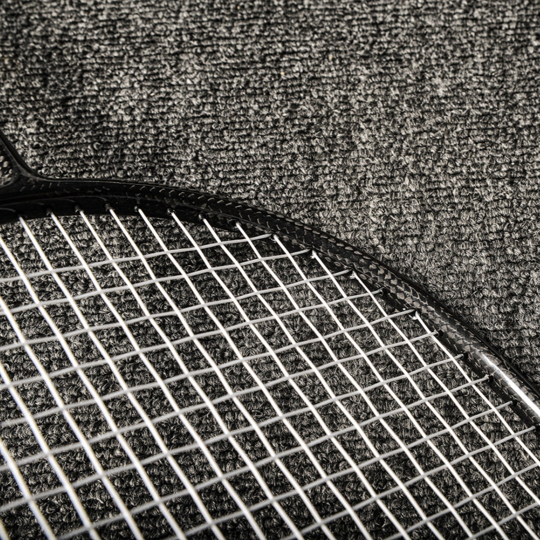 Close-up of a carbon fiber badminton racket featuring the textured frame and white strings, placed on a gray textured surface.