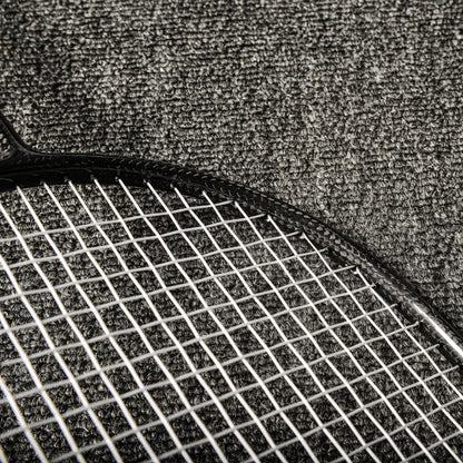 Close-up of a carbon fiber badminton racket featuring the textured frame and white strings, placed on a gray textured surface.