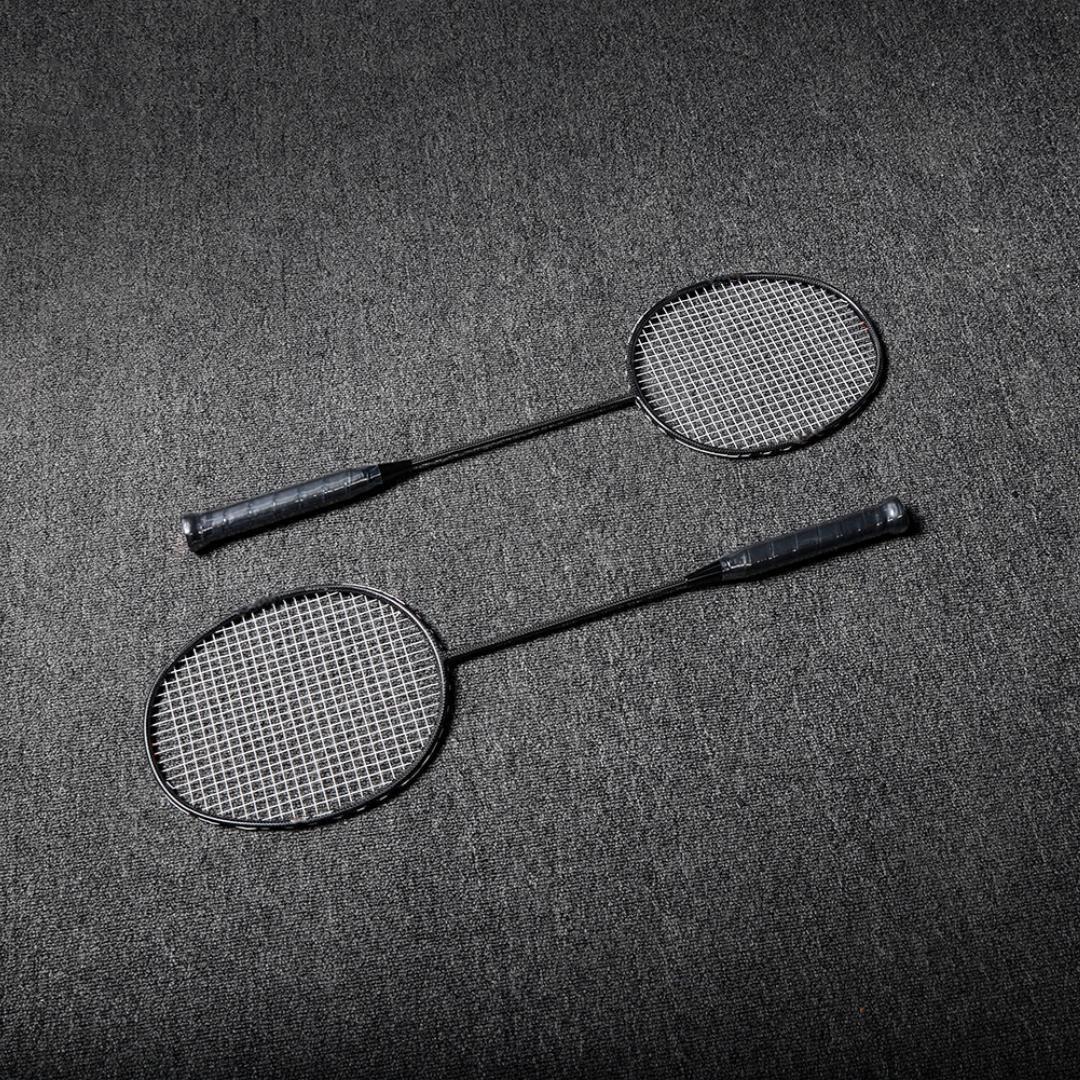 Close-up of two carbon fiber badminton rackets with black frames and white strings, laid on a gray textured surface.