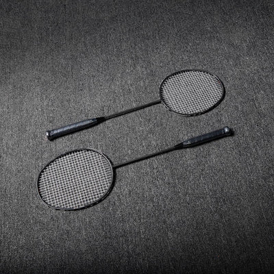 Close-up of two carbon fiber badminton rackets with black frames and white strings, laid on a gray textured surface.