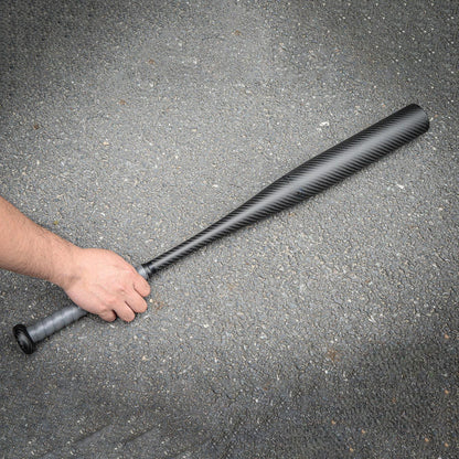 Hand holding a carbon fiber baseball bat with a distinct weave pattern, placed on an asphalt surface.