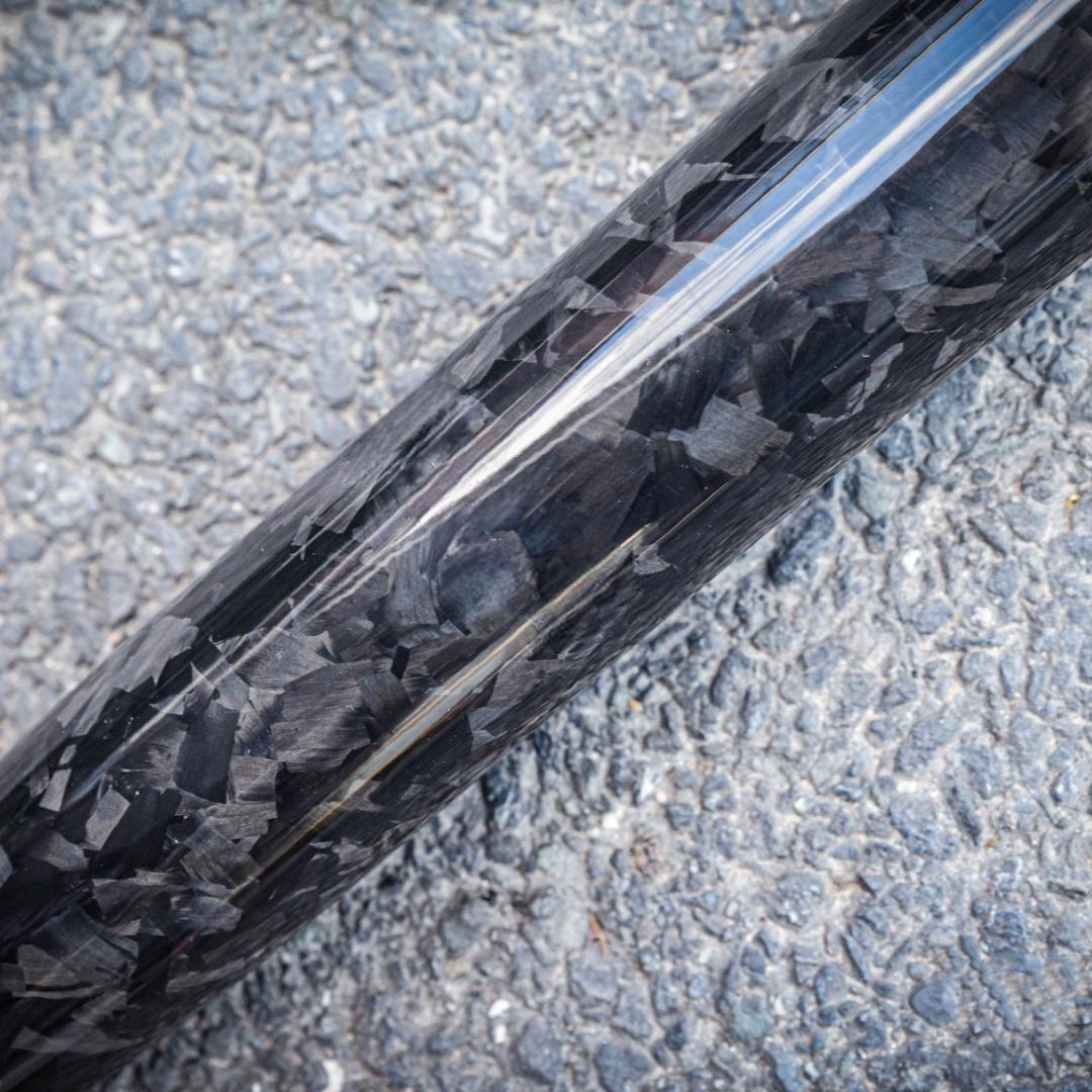 Close-up of a forged carbon fiber baseball bat with a distinct chopped carbon fiber pattern, placed on an asphalt surface.