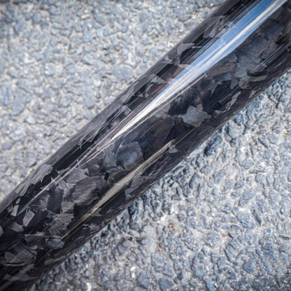 Close-up of a forged carbon fiber baseball bat with a distinct chopped carbon fiber pattern, placed on an asphalt surface.
