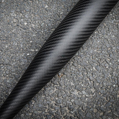 Close-up of a 3K carbon fiber baseball bat with a distinct weave pattern, placed on an asphalt surface.