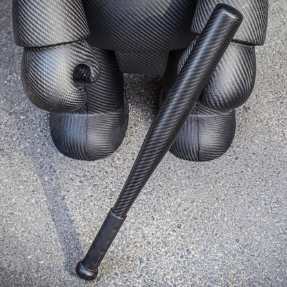 Close-up of a 3K carbon fiber baseball bat with a distinct weave pattern, positioned next to a large carbon fiber sculpture on an asphalt surface.