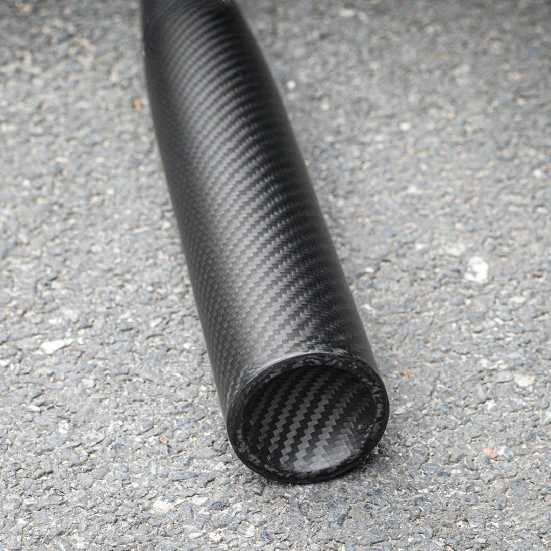 Close-up of a 3K carbon fiber baseball bat with a distinct weave pattern, showcasing the end and body detail, placed on an asphalt surface.