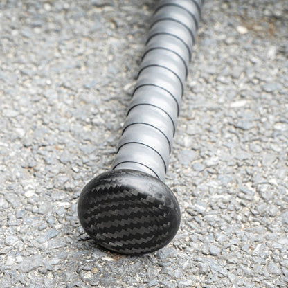 Close-up of the handle and end cap of a 3K carbon fiber baseball bat, showcasing the distinct carbon fiber weave pattern, placed on an asphalt surface.