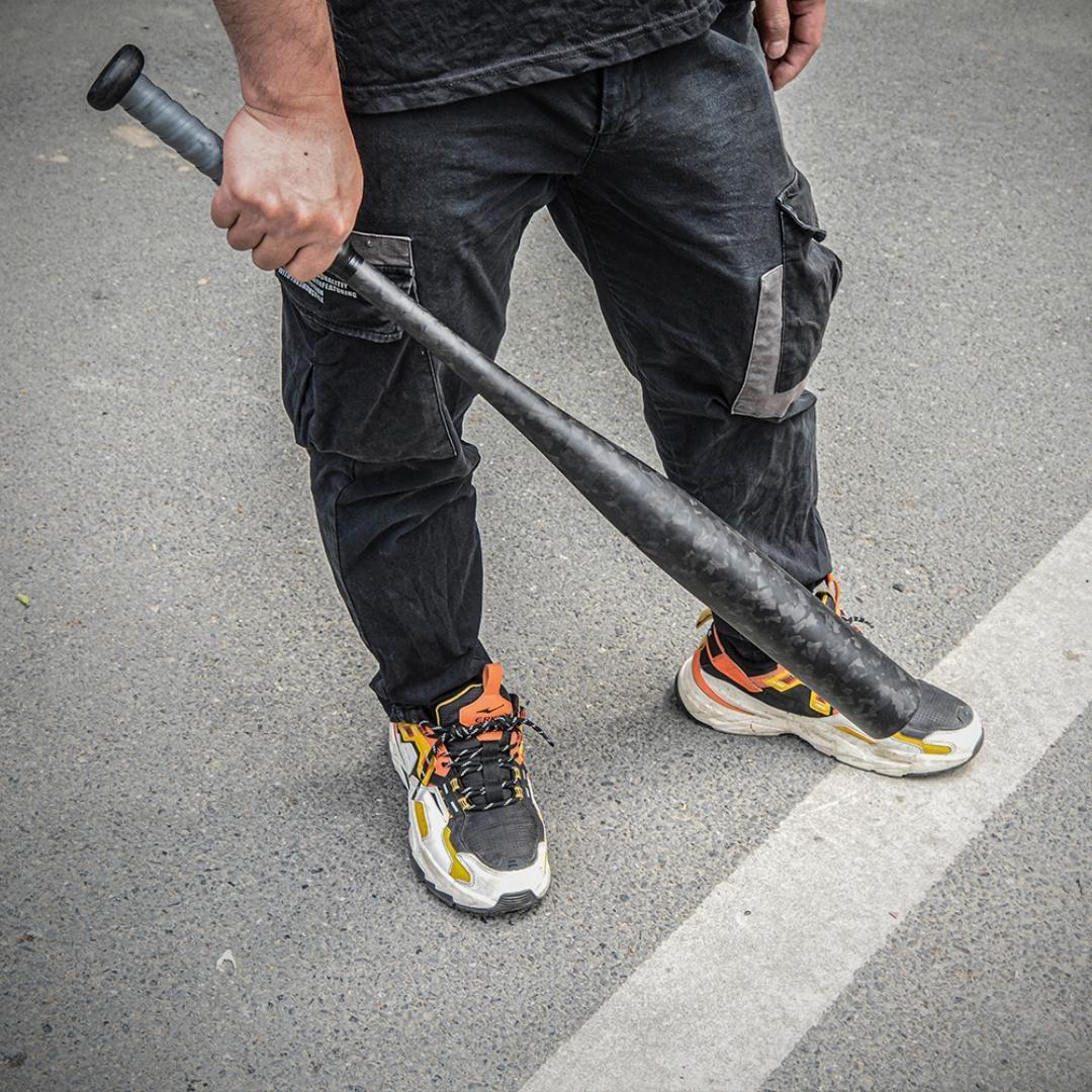 A person holding a forged carbon fiber baseball bat with a distinct chopped carbon fiber pattern, positioned on an asphalt surface.