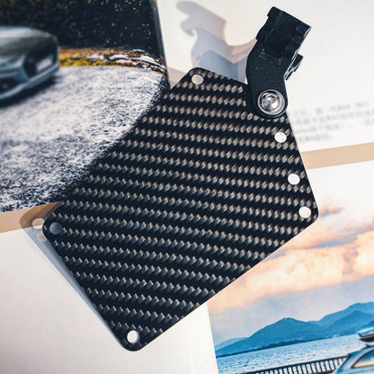 Close-up of a carbon fiber bicycle number plate with a distinct woven pattern, featuring a mounting clip, placed against a backdrop with automotive and landscape imagery.