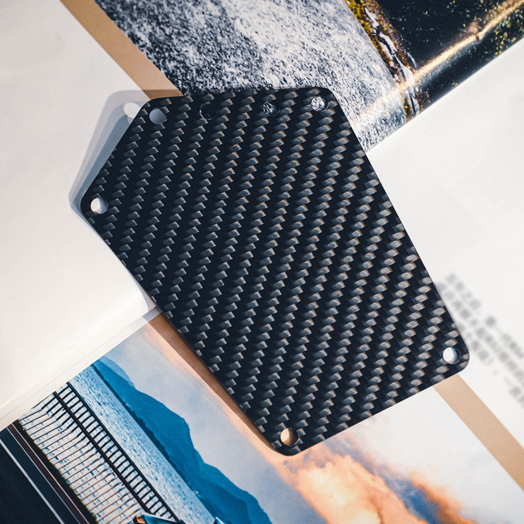 Close-up of a carbon fiber bicycle number plate with a distinct woven pattern and multiple mounting holes, placed on a backdrop featuring landscape and automotive imagery.