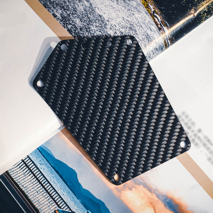 Close-up of a carbon fiber bicycle number plate with a distinct woven pattern and multiple mounting holes, placed on a backdrop featuring landscape and automotive imagery.