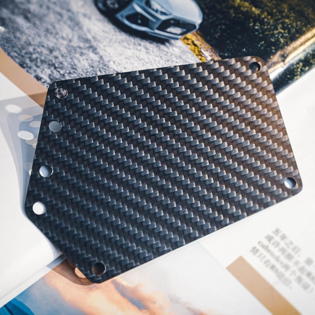 Close-up of a carbon fiber bicycle number plate showcasing its distinct woven pattern and multiple mounting holes, placed on a backdrop with automotive imagery.
