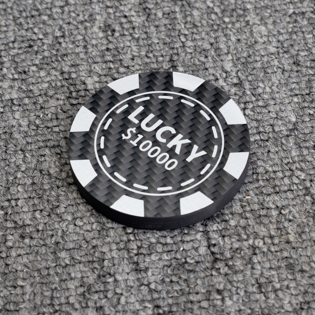 Close-up of a poker chip-style carbon fiber bottle opener with "LUCKY $10000" text, showcasing its carbon fiber texture, placed on a gray textured surface.