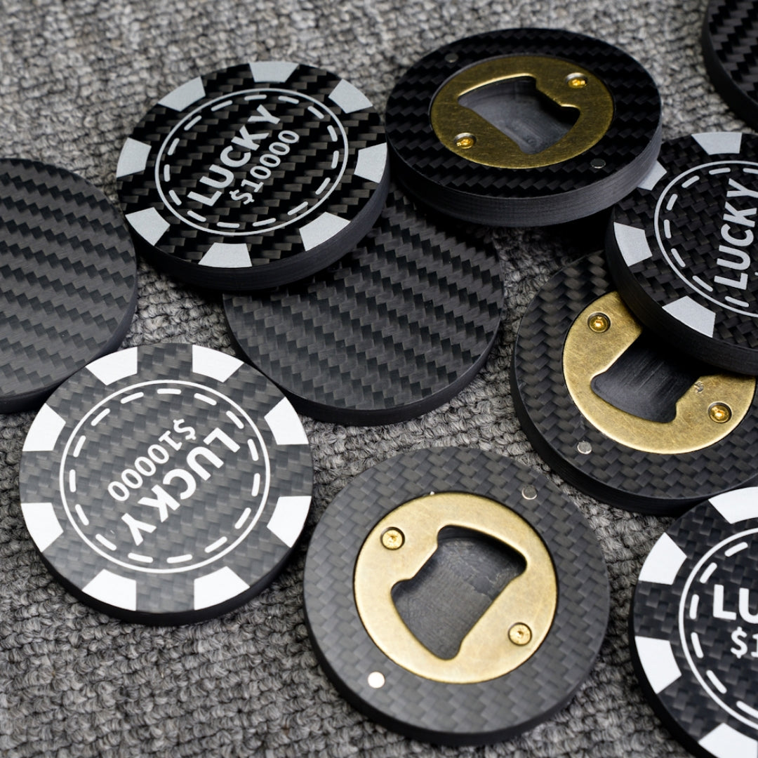 Close-up of multiple carbon fiber bottle openers, including poker chip-style ones with "LUCKY $10000" text and designs featuring brass bottle opener mechanisms, scattered on a gray textured surface.