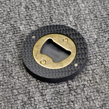 Close-up of a 3K carbon fiber bottle opener with a brass opener mechanism, showcasing its circular shape and distinct woven carbon fiber pattern, placed on a gray textured surface.