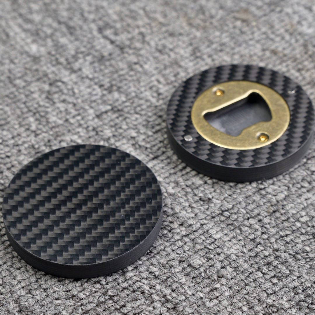 Close-up of two 3K carbon fiber bottle openers, showcasing their circular shape, distinct woven carbon fiber pattern, and brass opener mechanism, placed on a gray textured surface.
