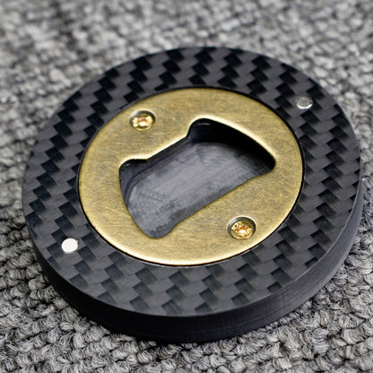 Close-up of a 3K carbon fiber bottle opener with a brass opener mechanism, showcasing its circular shape and distinct woven carbon fiber pattern, placed on a gray textured surface.
