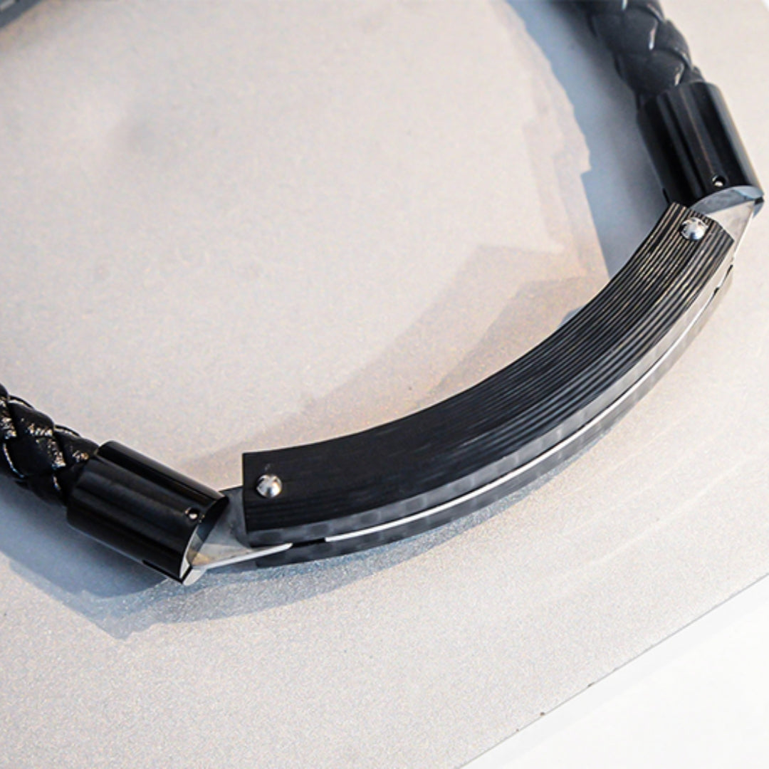 Close-up detail of a carbon fiber bracelet featuring a black braided strap, textured carbon fiber panel, and metallic accents, placed on a light-colored surface.