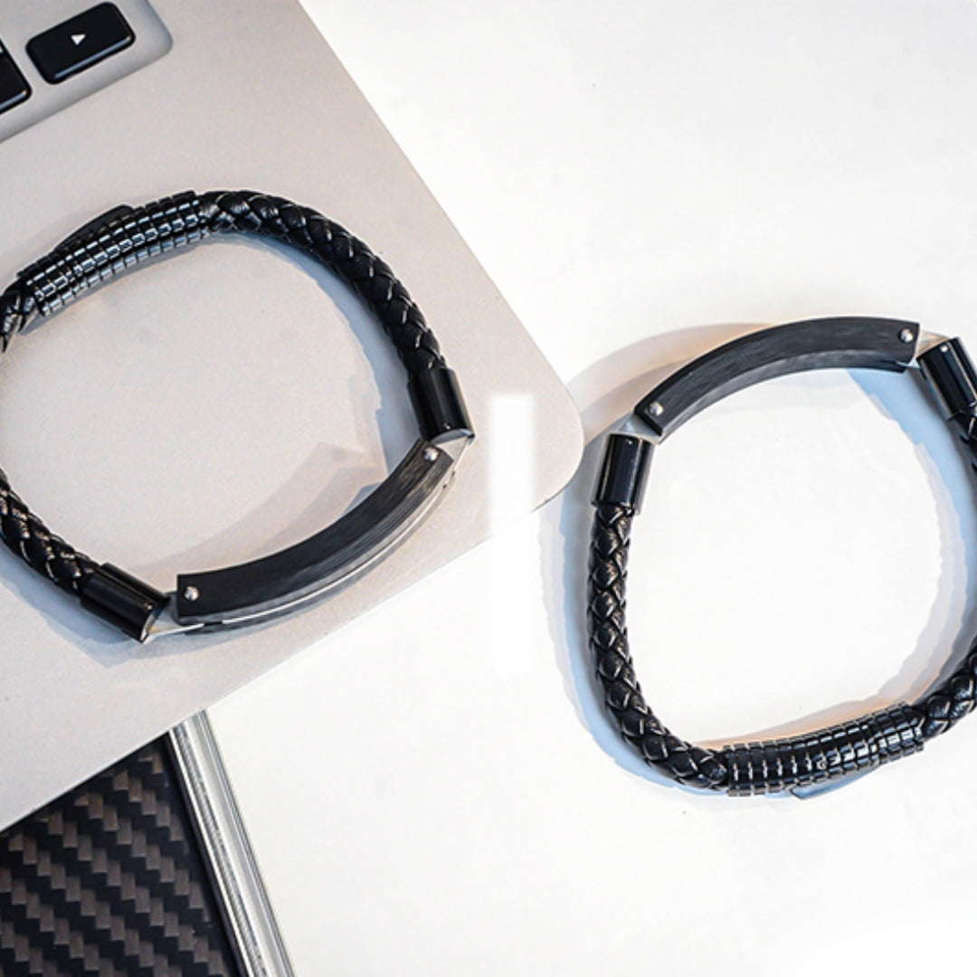 Close-up detail of two carbon fiber bracelets with black braided straps and textured carbon fiber accents, placed on a light-colored surface next to a laptop and a carbon fiber-patterned element.