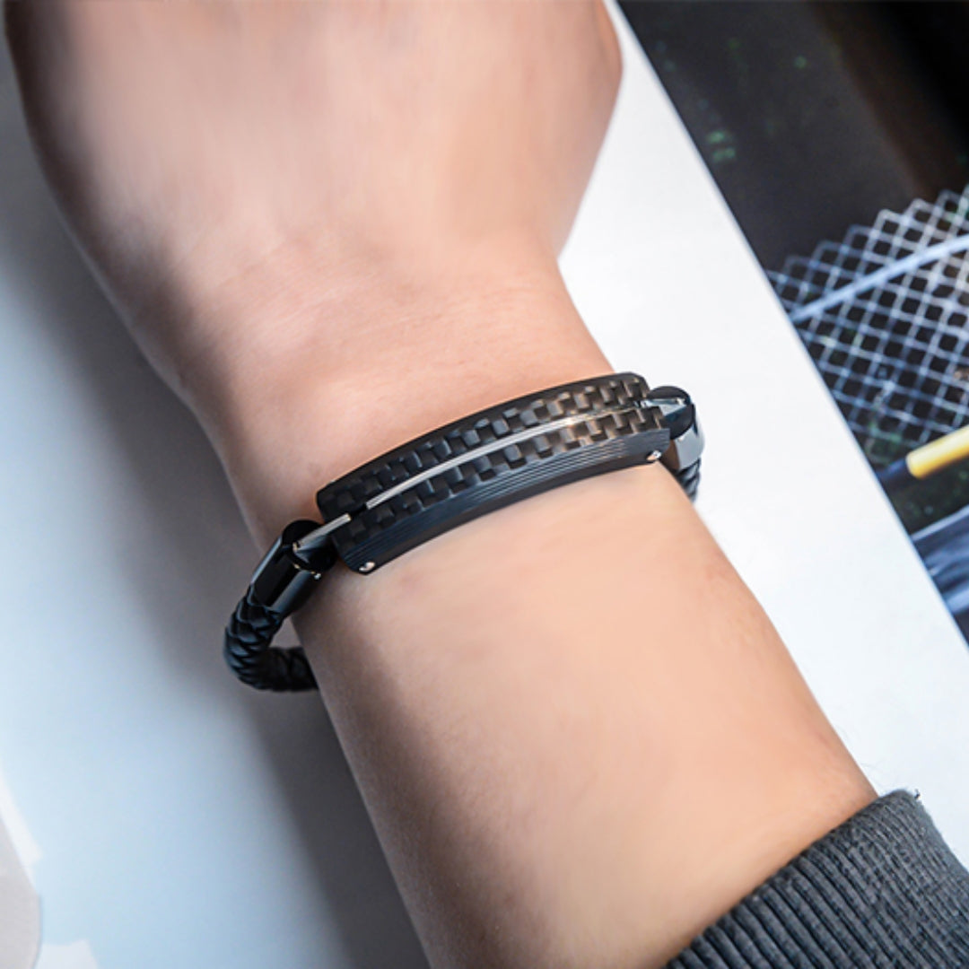 Close-up detail of a carbon fiber bracelet with a black braided strap and a textured carbon fiber accent, worn on a wrist against a light-colored surface with a patterned item in the background.
