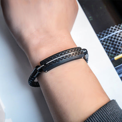 Close-up detail of a carbon fiber bracelet with a black braided strap and a textured carbon fiber accent, worn on a wrist against a light-colored surface with a patterned item in the background.
