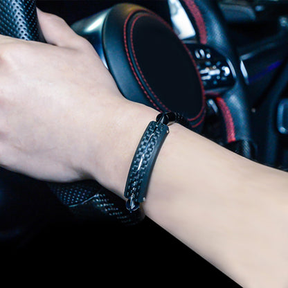 Close-up detail of a black carbon fiber bracelet worn on a wrist, with a car steering wheel featuring red stitching in the background.
