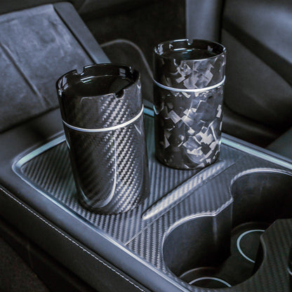 Close-up of two carbon fiber car ashtrays—one with a 3K carbon fiber pattern, the other with a forged carbon fiber pattern—placed on a carbon fiber-trimmed center console in a vehicle.