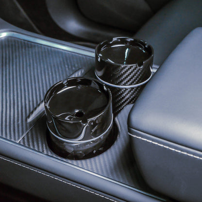 Close-up of two car ashtrays—one with a 3K carbon fiber pattern, the other with a forged carbon fiber pattern—placed in the cup holders of a car's center console featuring carbon fiber trim.
