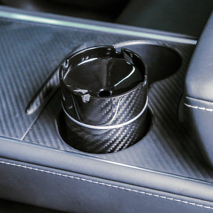 Close-up detail of a 3K carbon fiber car ashtray with a woven pattern, placed in a cup holder of a car's center console featuring carbon fiber trim and leather upholstery.