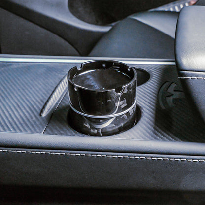 Close-up of a forged carbon fiber car ashtray with a distinct fragmented pattern, placed in the cup holder of a car's center console featuring carbon fiber trim and leather upholstery.