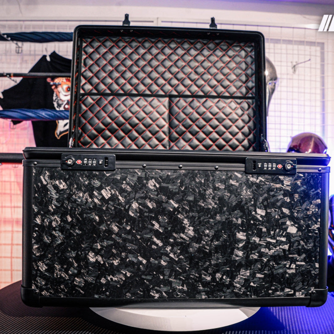 Close-up detail of an open large car storage trunk made of forged carbon fiber with a chopped texture, featuring a quilted interior with red stitching and combination locks, displayed in a stylishly lit space with decorative background elements.