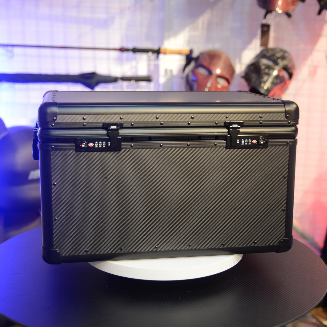 Close-up detail of a closed large 3K carbon fiber car storage trunk with combination locks, displayed on a round table against a background featuring decorative masks and fishing gear.
