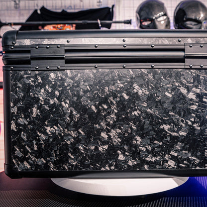 Close-up detail of the back side of a large forged carbon fiber car storage trunk, showcasing the chopped carbon fiber pattern, metallic rivets, and structural details, with decorative masks visible in the background.