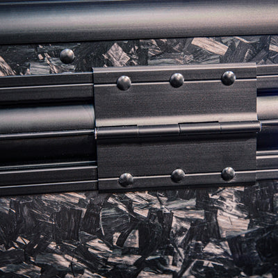 Close-up detail of the back side of a large forged carbon fiber car storage trunk, highlighting the chopped carbon fiber pattern and a metal hinge with rivets.