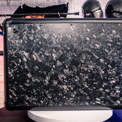 Close-up detail of the bottom part of a large forged carbon fiber car storage trunk, showcasing the chopped carbon fiber pattern and metal edges, with decorative masks visible in the background.