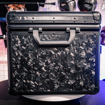 Close-up side view of a large forged carbon fiber car storage trunk, showcasing the chopped carbon fiber pattern and black handle, with decorative masks visible in the background.