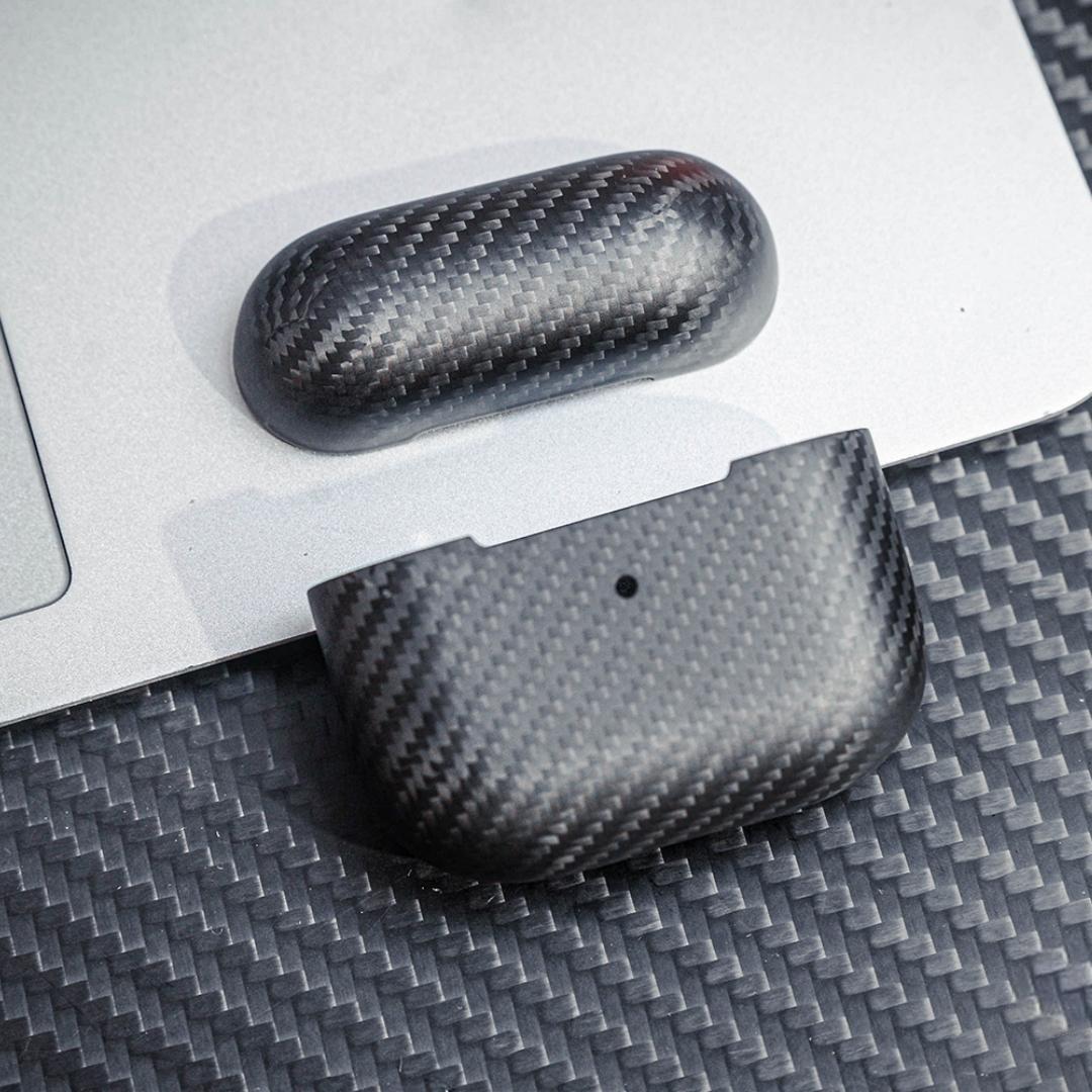 Close-up of 3K carbon fiber AirPods cases showcasing the distinct weave pattern, displayed on a surface with matching carbon fiber texture and a metallic background.