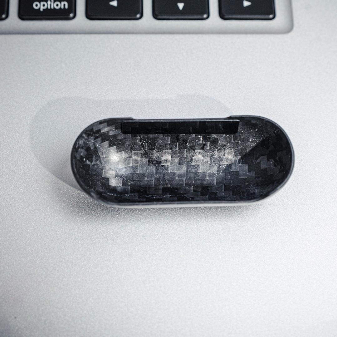 Close-up of the interior of the top half of a 3K carbon fiber AirPods case, showcasing the distinct weave pattern, placed on a metallic surface with a keyboard in the background.